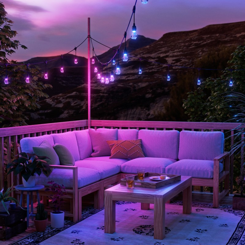 Outdoor string lights in purple and blue hues hanging from a pole on a wooden patio with a scenic mountain view. Coffee table with a tray of two drinks and a plate. A corner bench chair with surrounding greenery and plants.